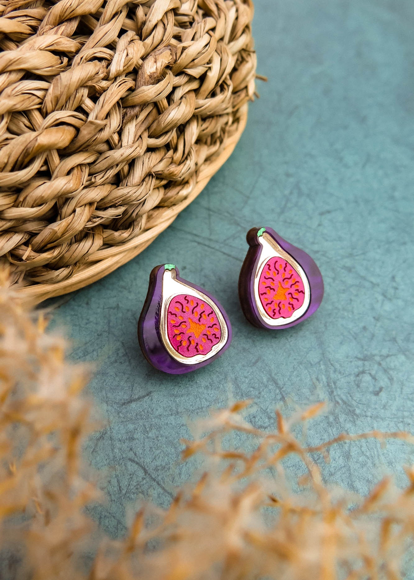 A pair of fig-shaped, hand-painted earrings with purple and pink detailing are displayed on a green surface next to a woven basket. Blurred beige plant stems are visible in the foreground.