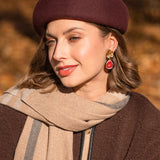 A woman wearing a maroon beret, beige and brown scarf, and matching coat smiles softly outdoors. She has light makeup and wears handcrafted Fig Earrings with gold mirror leaves. The background is blurred with autumn colors.