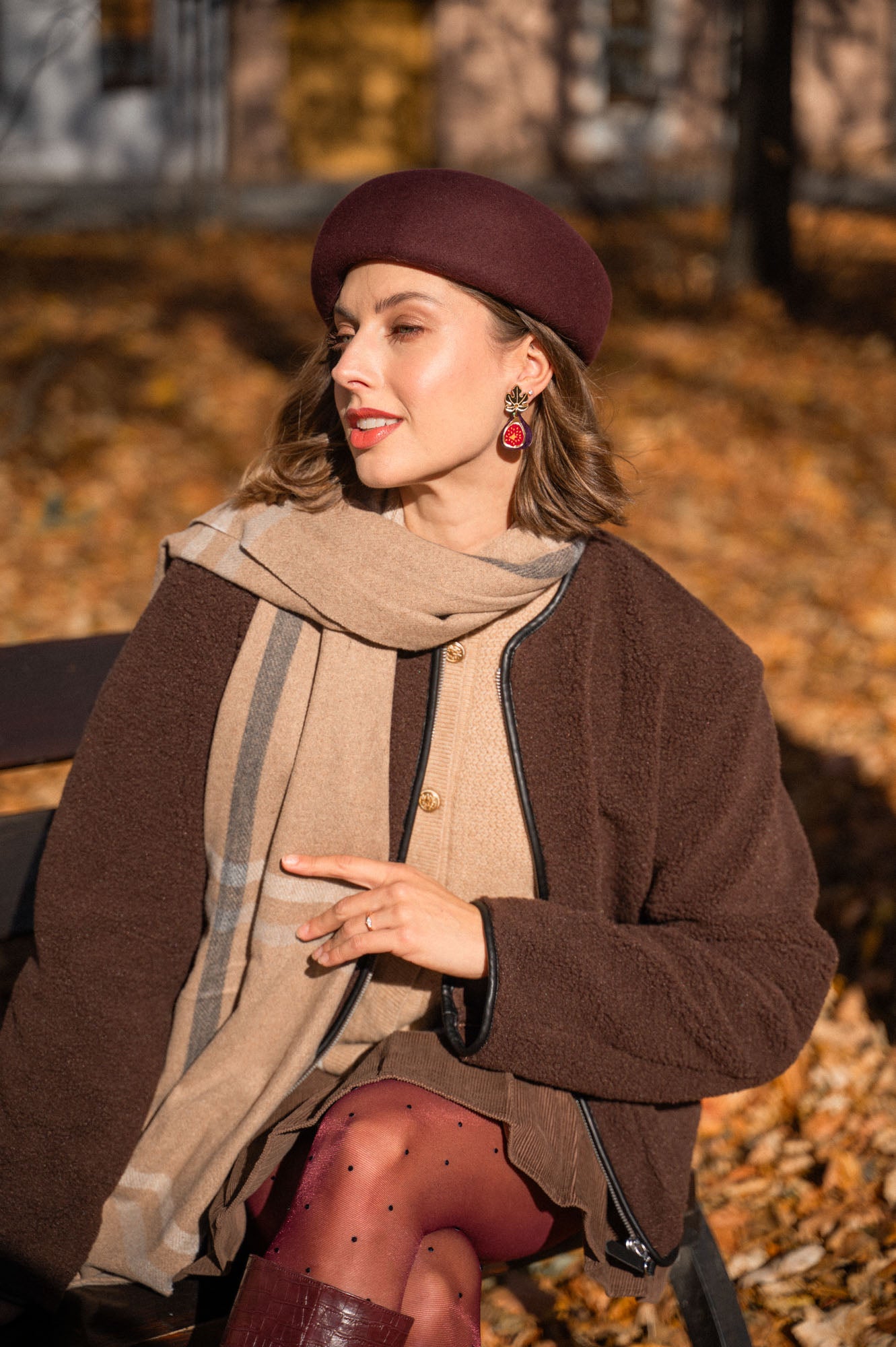A woman wearing a brown beret, a brown coat, tan scarf, and handcrafted earrings sits outside on a bench surrounded by autumn leaves, looking to the side and smiling softly in the sunlight.