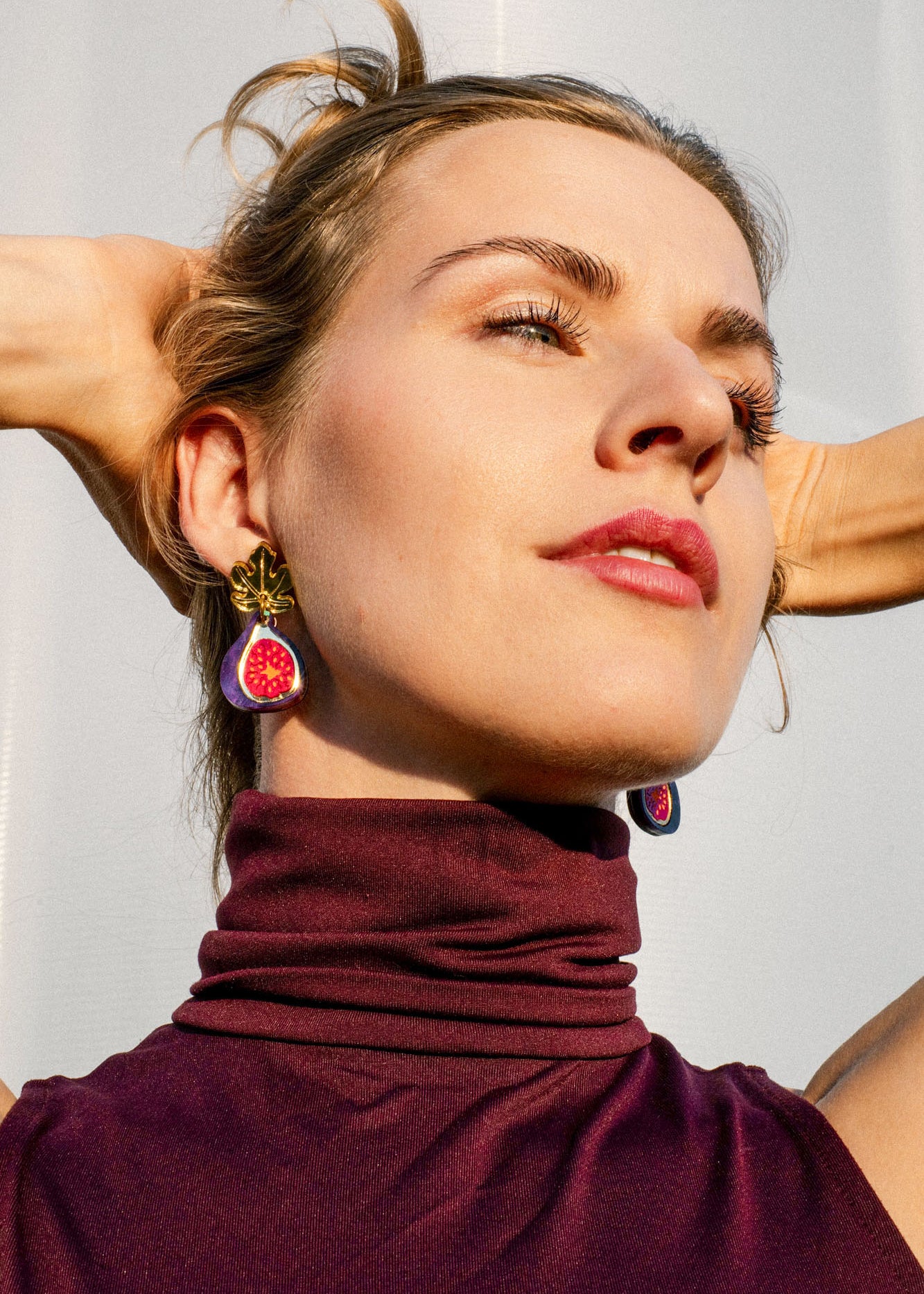 Woman wearing a sleeveless maroon turtleneck top and handcrafted fig-shaped earrings with delicate gold mirror leaves poses with arms raised behind her head, looking upward against a light background.