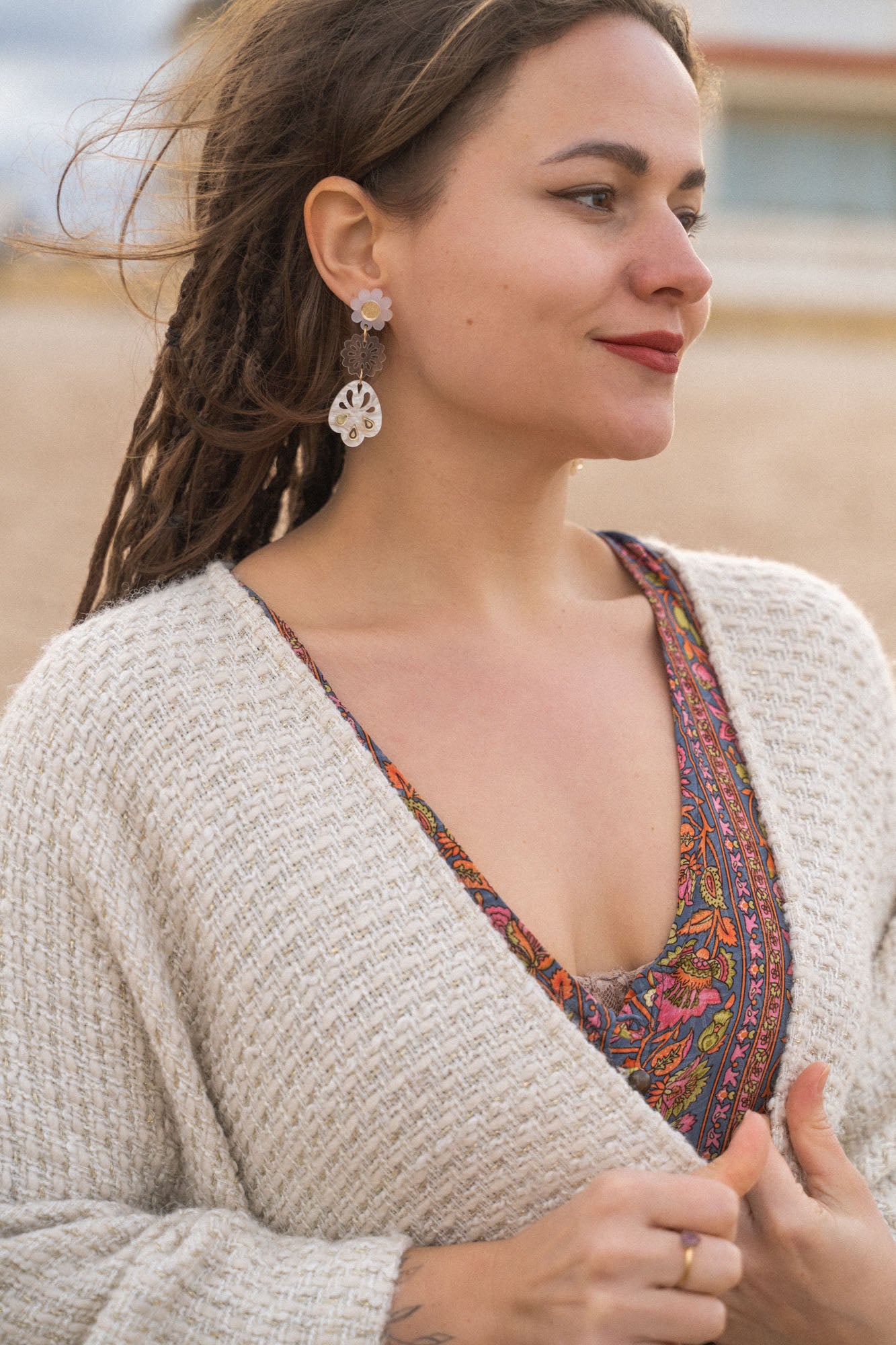 A woman with long braided hair stands outdoors, wearing a patterned dress and a cream knit sweater. She has handcrafted boho earrings and is smiling softly, looking to the side. The background appears to be a beach or sandy area.
