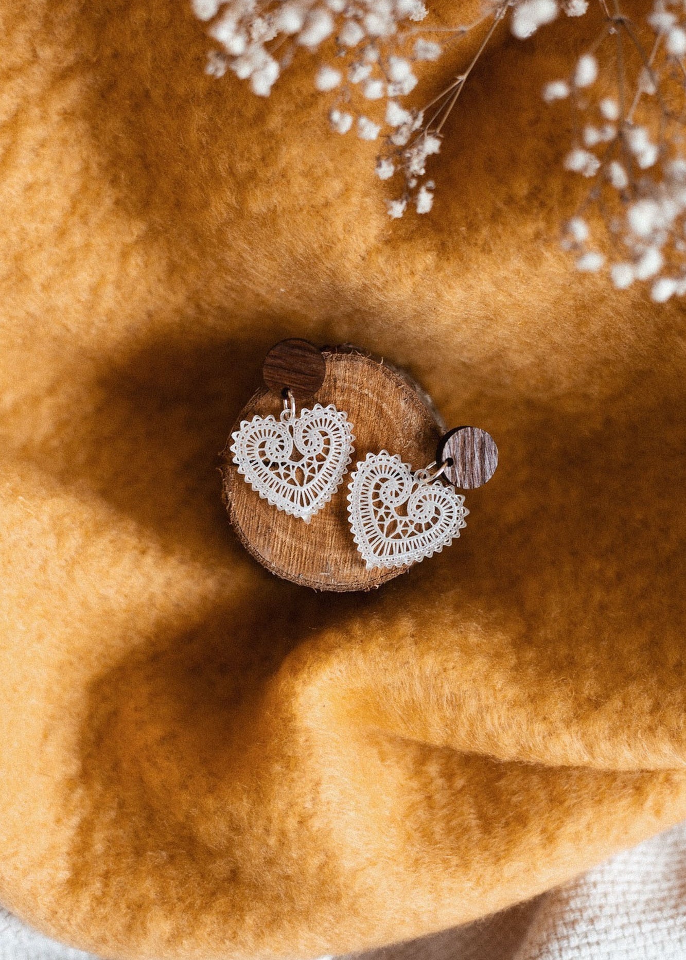 A pair of Folk Heart Earrings, showcasing lightweight elegance and delicate, white lace heart-shaped design, displayed on a small wooden disk atop soft golden-yellow fabric with white babys breath flowers in the background.