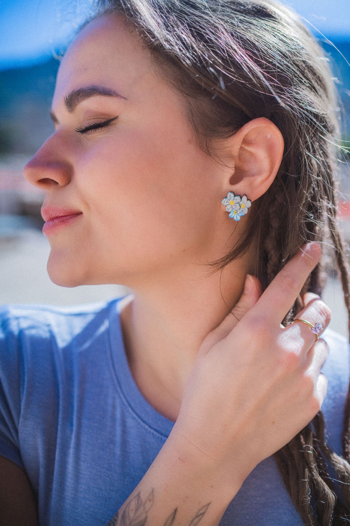 A woman with braided hair and Forget Me Not Studs smiles softly with her eyes closed, gently touching her hair. She wears a light blue shirt and stands outdoors in bright sunlight, showcasing the floral earrings as delicate wearable art.