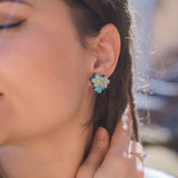 Close-up of a woman’s face, eyes closed, wearing Forget Me Not Studs—delicate floral earrings as wearable art. She touches her braided hair with one hand while sunlight softly illuminates her skin.