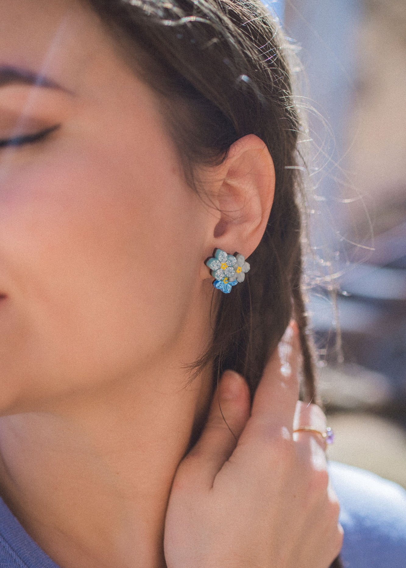 Close-up of a woman’s face, eyes closed, wearing Forget Me Not Studs—delicate floral earrings as wearable art. She touches her braided hair with one hand while sunlight softly illuminates her skin.