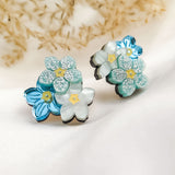 A pair of decorative Forget Me Not Studs featuring layered blue and white flowers with glittery petals, displayed on a soft white fabric with dried plants in the background—delicate floral earrings that are true wearable art.