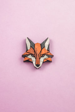 A geometric Fox Pin brooch, crafted from recycled wood in orange, black, and white hues, is displayed on a pink background—perfect for an eco-conscious style.
