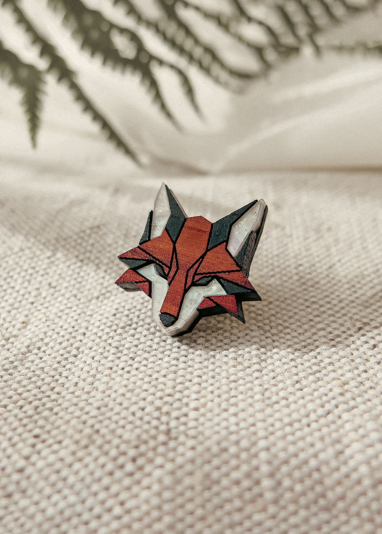 A geometric, handcrafted fox pin made from recycled wood rests on textured beige fabric, with blurred green fern leaves in the background.