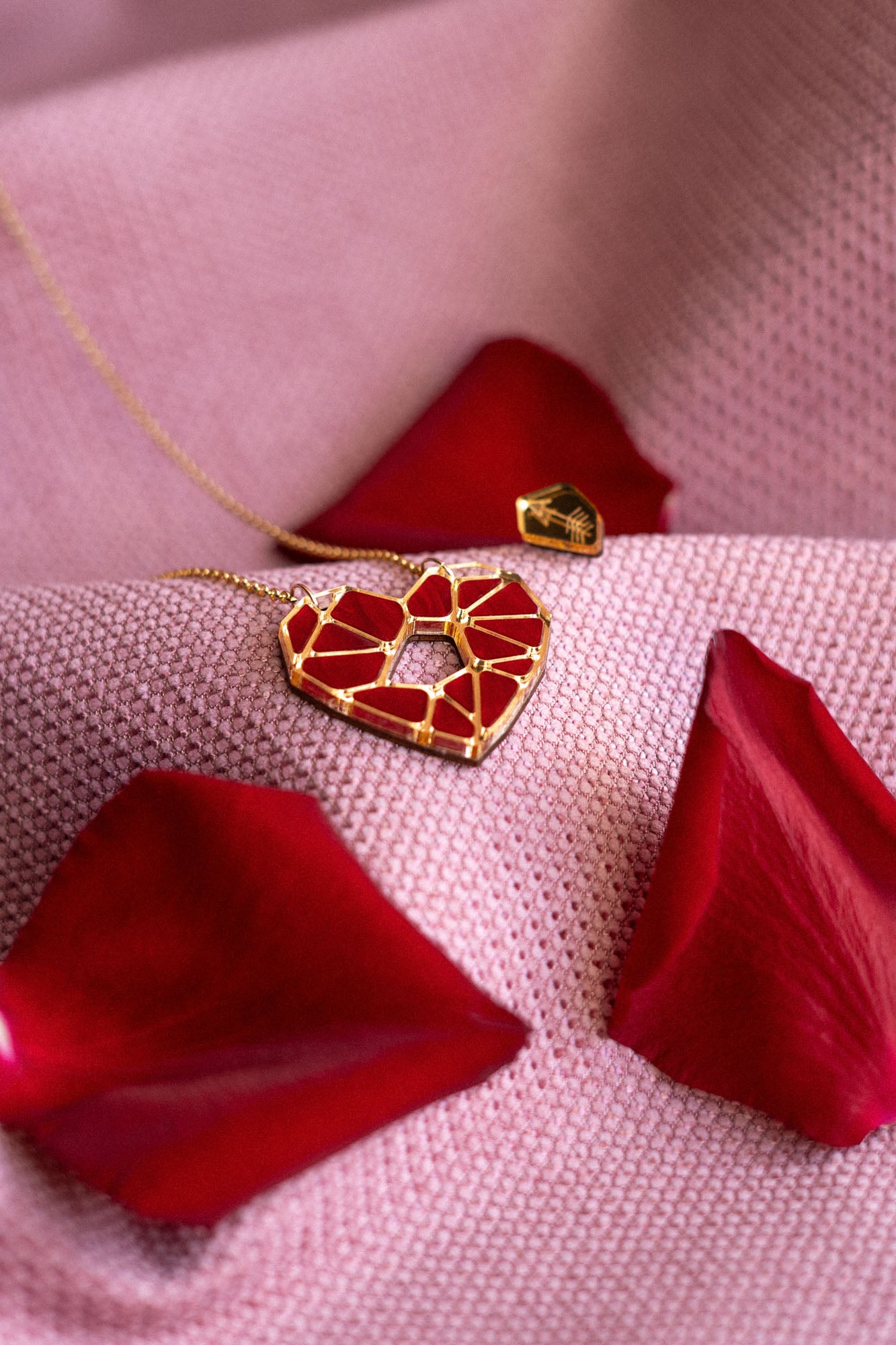 A handcrafted gold Fragment of Love Necklace with a geometric red heart pendant rests on soft pink fabric, surrounded by scattered red rose petals.