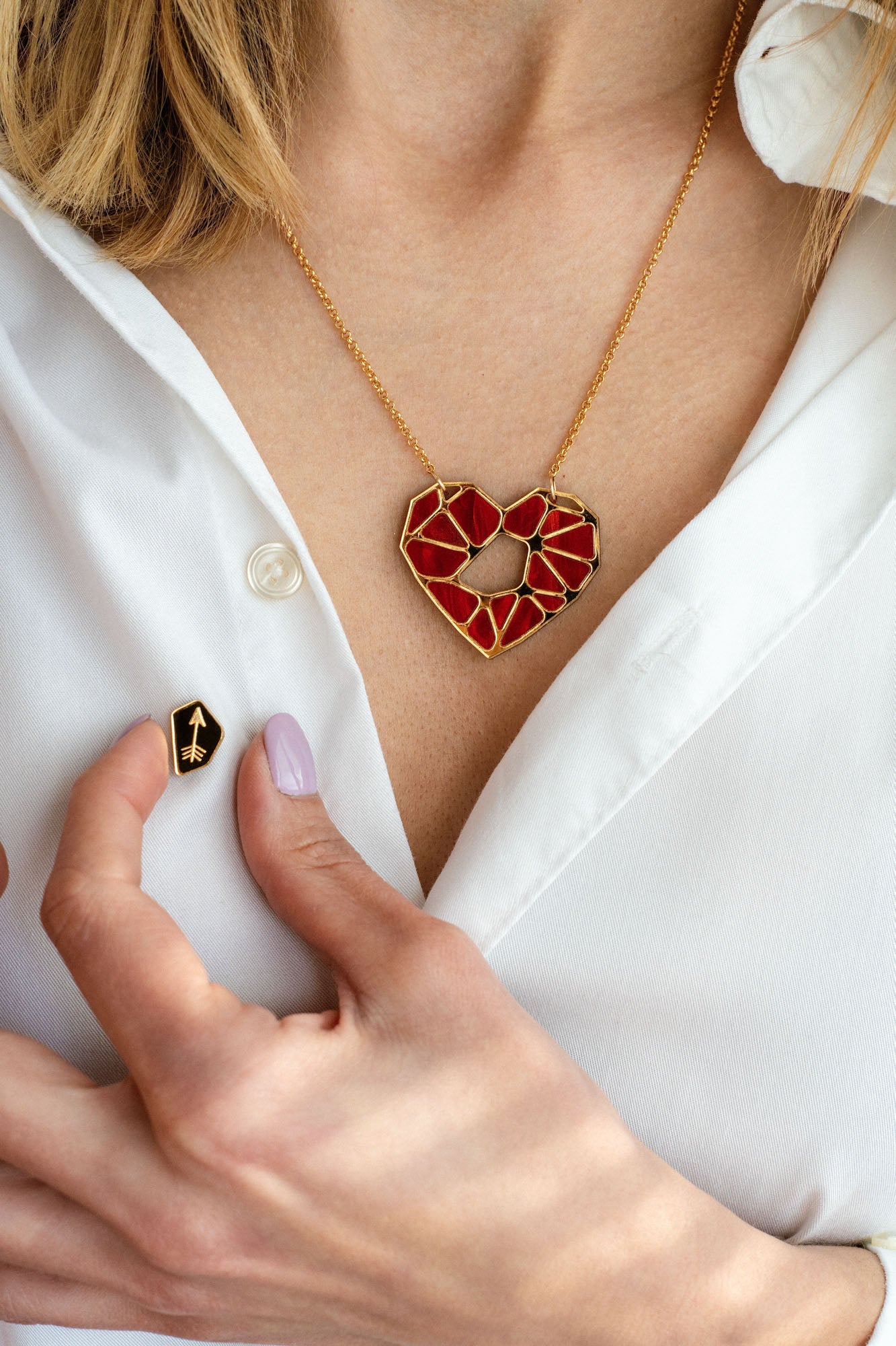 A person wearing a white shirt displays the handcrafted Fragment of Love Necklace featuring a red, geometric heart pendant and holds a small black pin with a gold arrow design. Only the lower face and hand are visible.