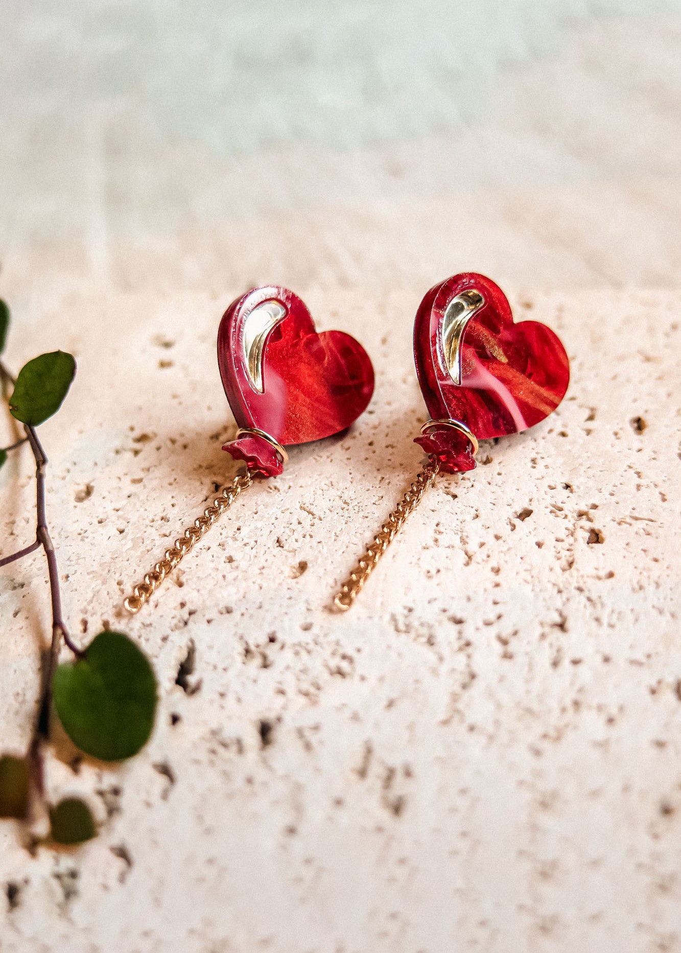 A pair of Heart Balloon Studs with a playful, vibrant style—red heart-shaped earrings featuring gold detailing and delicate chains, displayed on a textured beige surface beside a small green leafy plant.