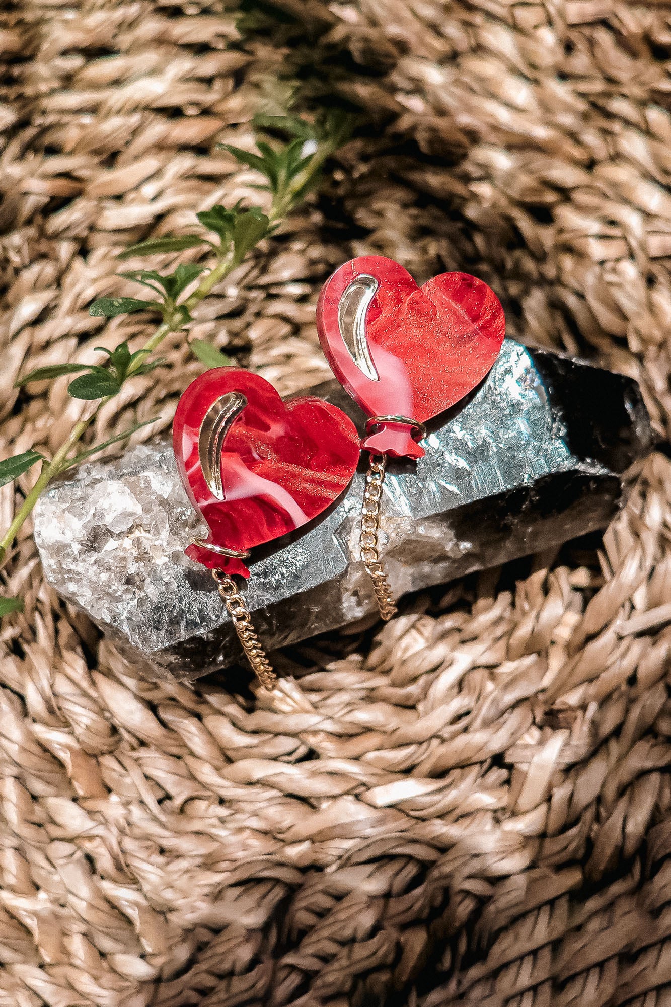 Two red Heart Balloon Studs with gold accents and playful dangling chains are displayed on a large crystal, resting on a woven, natural fiber surface with a small green plant nearby.