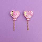 A pair of Heart Balloon Studs—pink, heart-shaped earrings with gold crescent accents and dangling gold chains—adds vibrant style, displayed on a purple background.