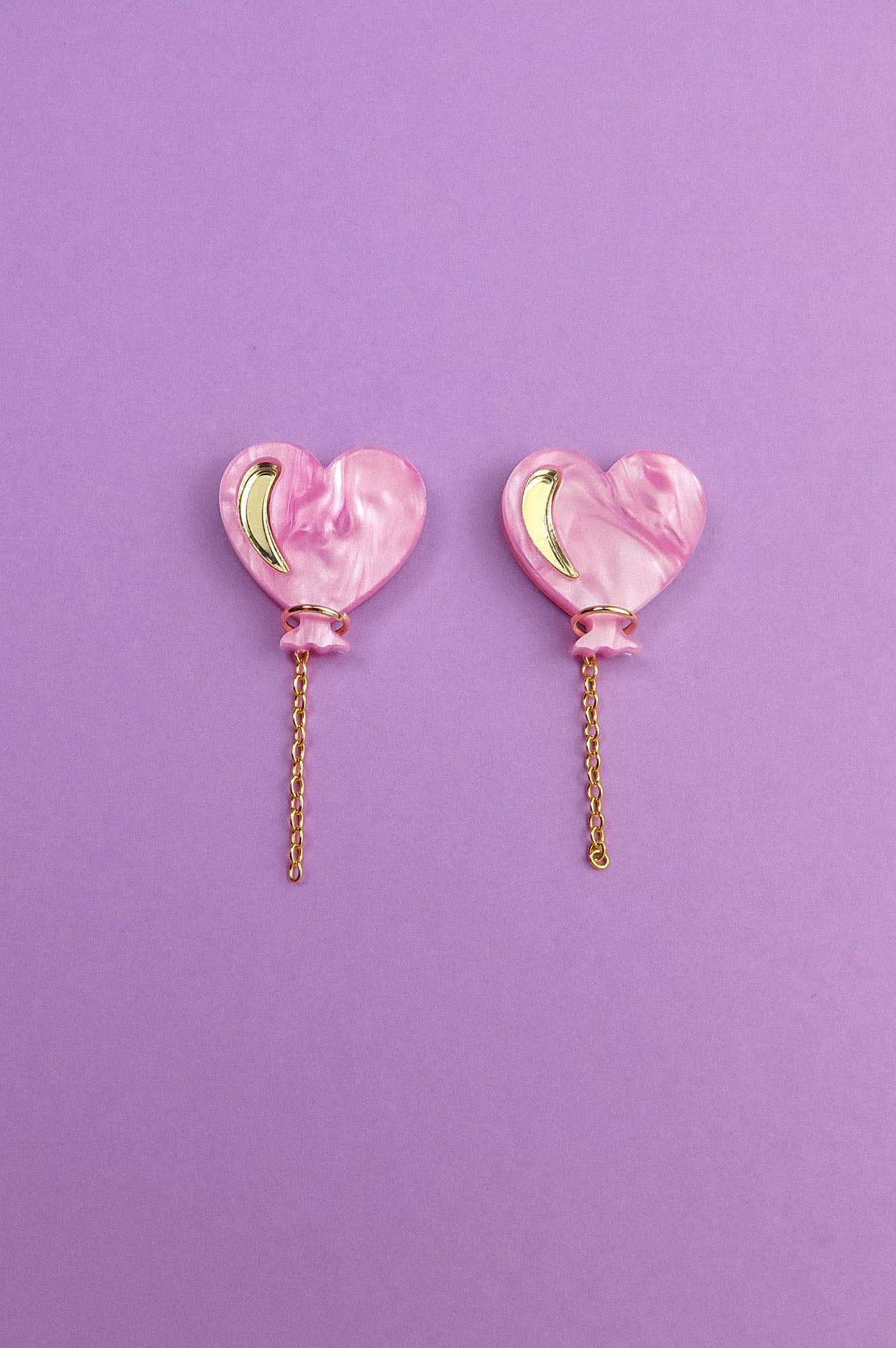 A pair of Heart Balloon Studs—pink, heart-shaped earrings with gold crescent accents and dangling gold chains—adds vibrant style, displayed on a purple background.