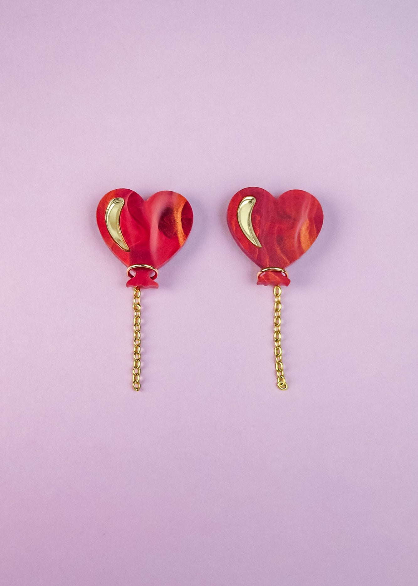 Two red marbled Heart Balloon Studs with small gold crescent details and playful gold chains hanging from the bottom, placed side by side on a light purple background.
