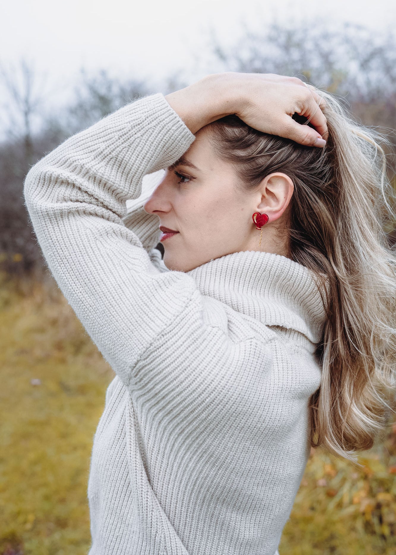 A woman in a cream-colored sweater stands outdoors, facing sideways with one hand holding back her long blonde hair. She wears playful Heart Balloon Studs, adding a romantic jewelry touch to her look. The background is blurry with trees and grass.