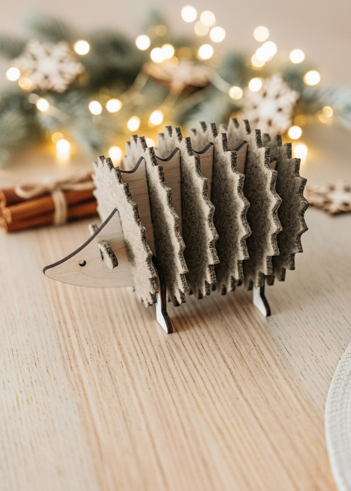 A wooden hedgehog figurine, perfect for home decor, sits on a light wooden table. In the blurred background, lights, fir branches, and cinnamon sticks create a festive, cozy atmosphere.