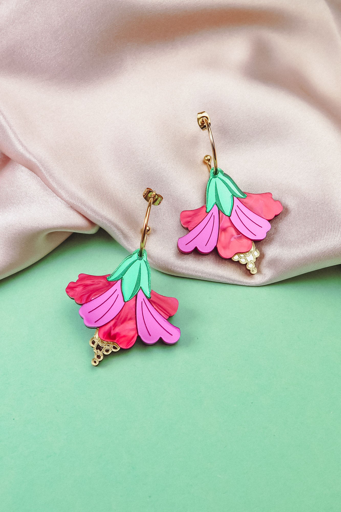 Handcrafted Hibiscus Earrings in a tropical statement style, featuring colorful pink and red petals, green leaves, and gold accents, are displayed on soft pink fabric and a mint green background.
