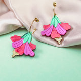 Two colorful Hibiscus Earrings with gold hoops and pink, purple, and green petal designs rest on light pink satin fabric and a mint green surface, making a handcrafted tropical statement.
