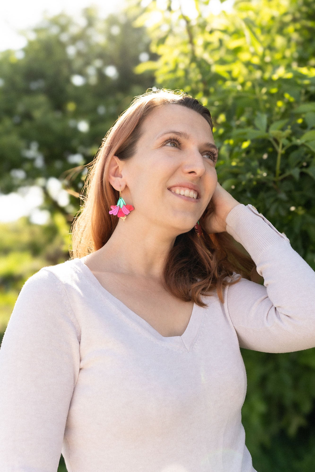 A woman with light brown hair smiles and looks upward, standing outdoors in sunlight. She wears a light long-sleeve top and handcrafted Hibiscus Earrings, with greenery and trees in the background.