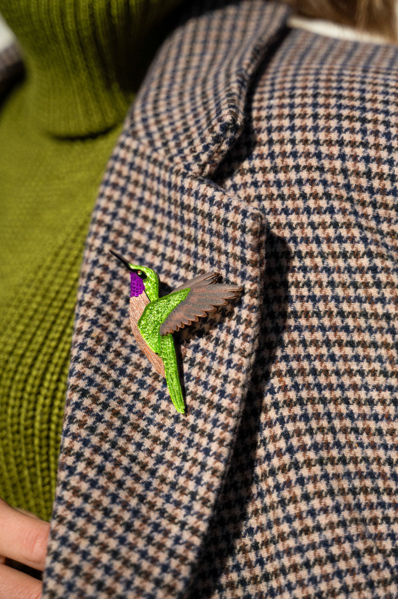 A close-up of a handcrafted hummingbird brooch, made from premium materials, pinned to a houndstooth-patterned coat with a green turtleneck sweater visible underneath.