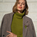 A woman wearing a green turtleneck sweater and a brown plaid coat stands against a light-colored brick wall, smiling slightly with her hand resting on her coat adorned with a handcrafted Hummingbird Brooch.