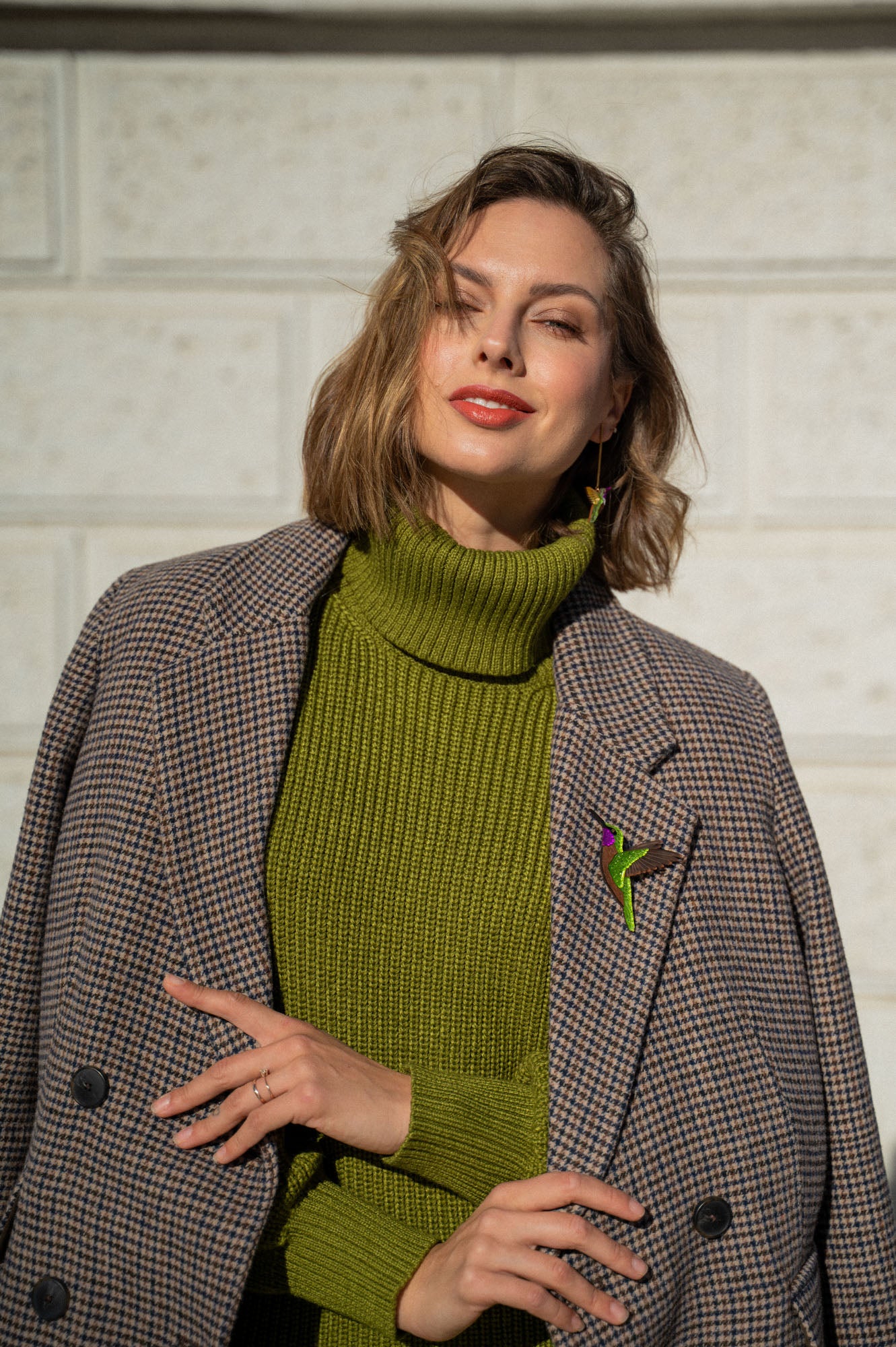 A woman wearing a green turtleneck sweater and a brown plaid coat stands against a light-colored brick wall, smiling slightly with her hand resting on her coat adorned with a handcrafted Hummingbird Brooch.
