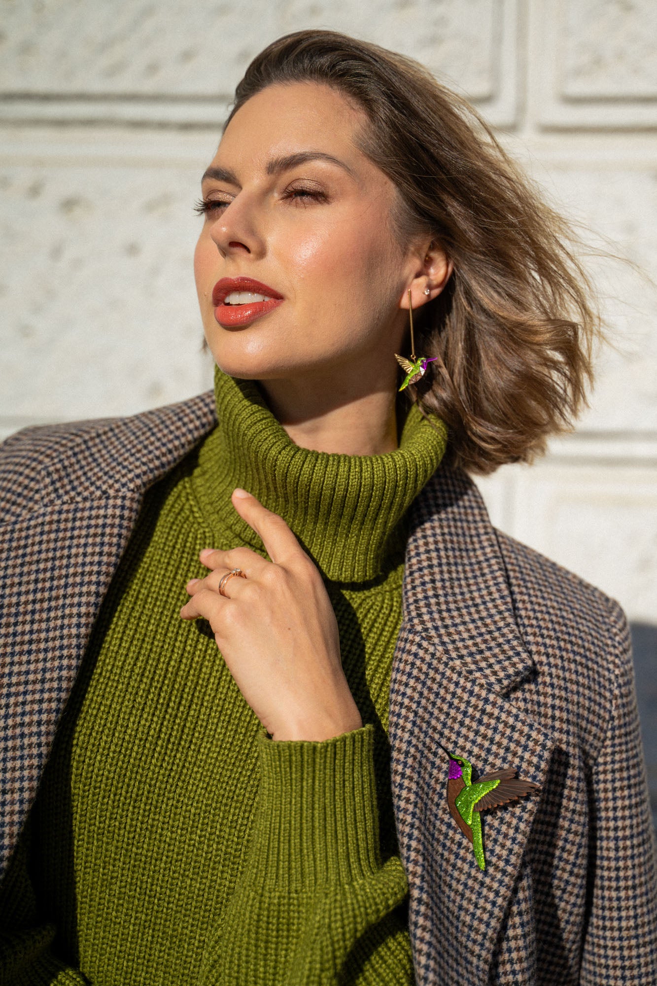 Woman wearing a green turtleneck sweater and plaid coat, accessorized with handcrafted Hummingbird Earrings and a brooch, poses outdoors against a light textured wall, touching her collar and looking off to the side.