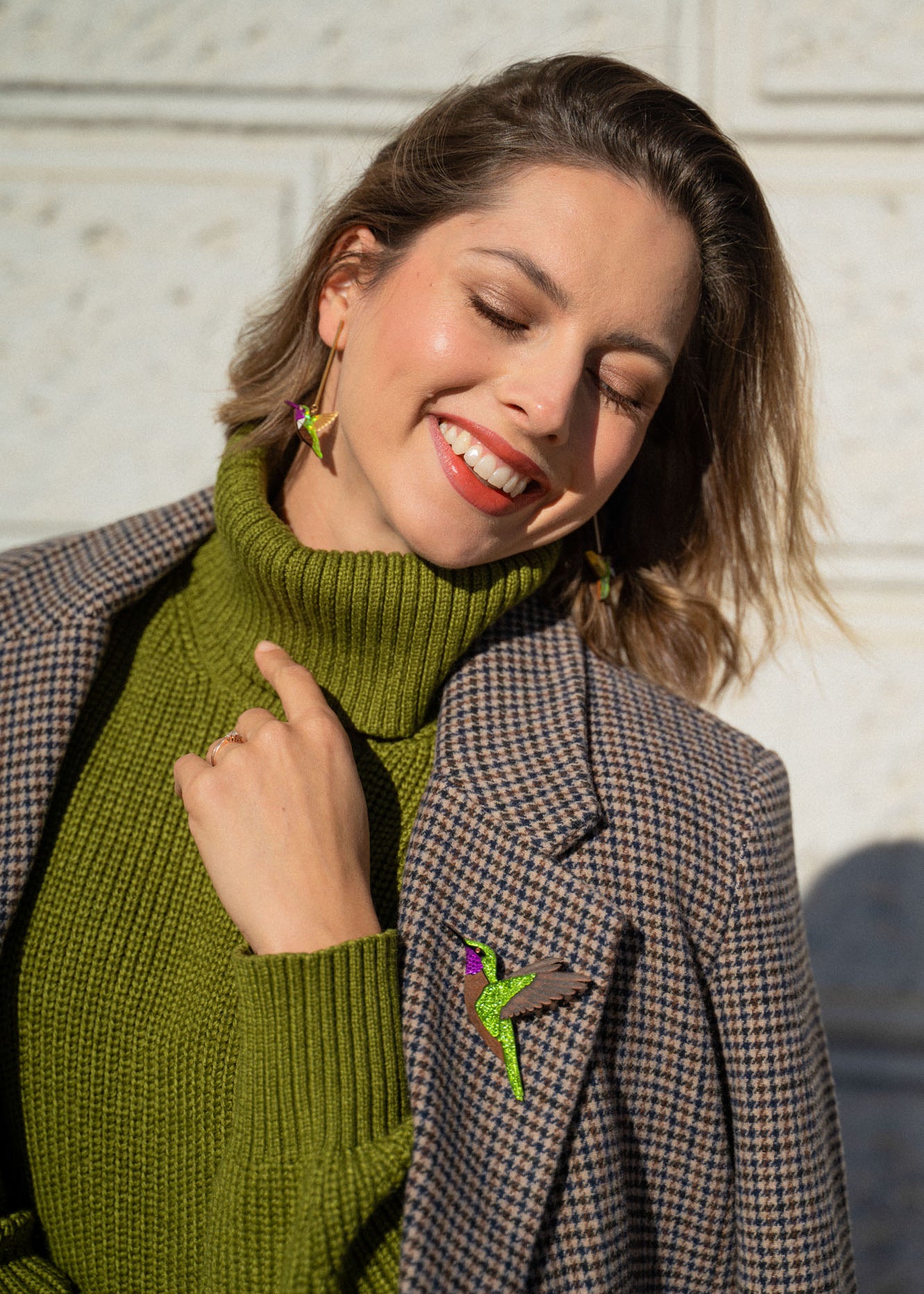 A smiling woman in a green turtleneck and plaid blazer stands in sunlight, eyes closed and head tilted. She wears bird-shaped earrings and a handcrafted Hummingbird Brooch, all made from premium materials.