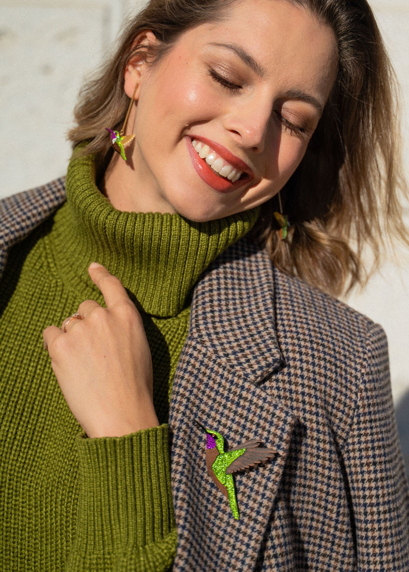 A smiling woman in a green turtleneck and plaid blazer stands in sunlight, eyes closed and head tilted. She wears bird-shaped earrings and a handcrafted Hummingbird Brooch, all made from premium materials.