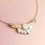 A handcrafted gold Tulip Necklace with a delicate chain and a curved bar pendant engraved with small symbols, featuring a cloud-shaped white charm, displayed on a pink background for added romantic charm.