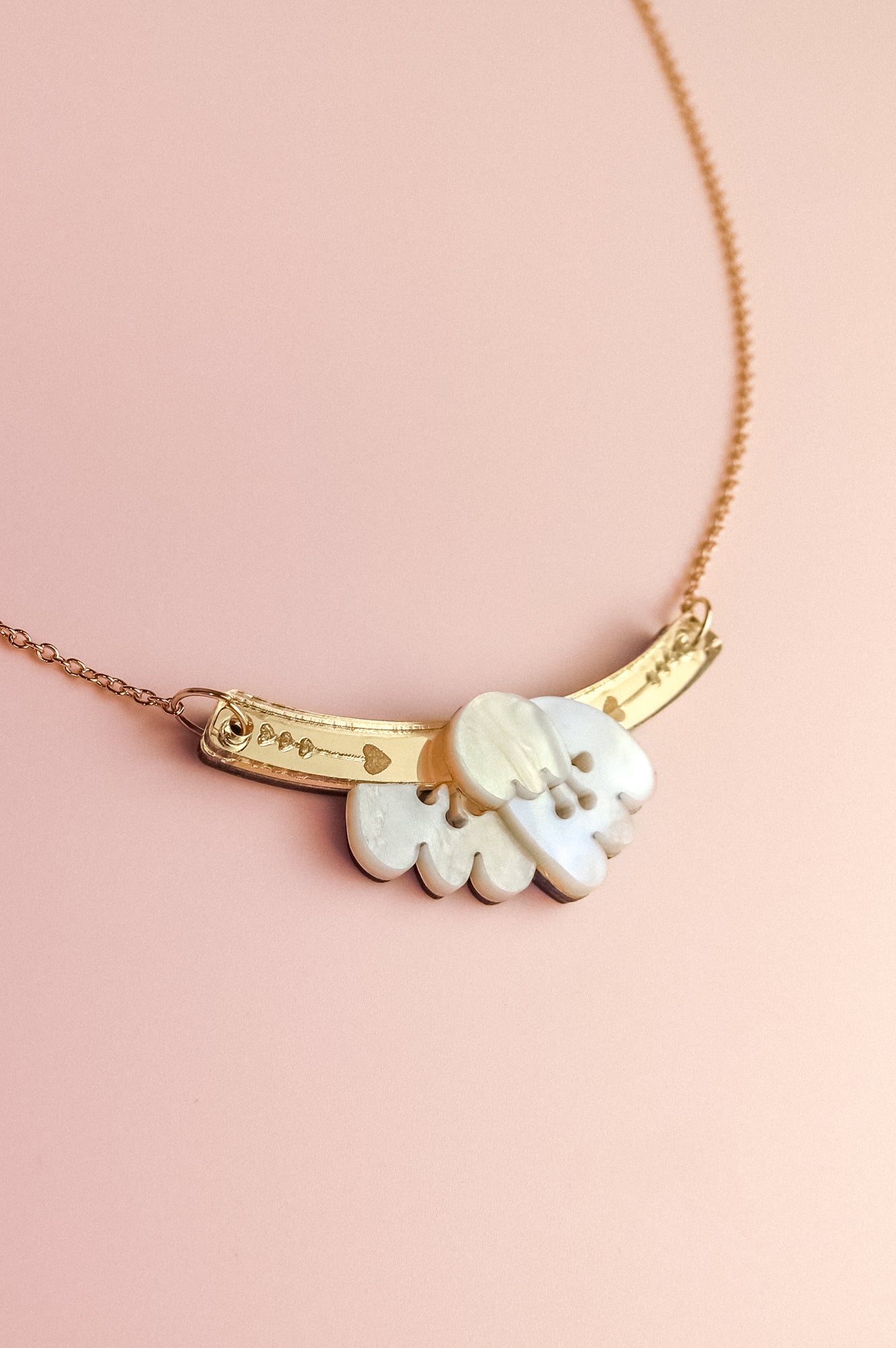 A handcrafted gold Tulip Necklace with a delicate chain and a curved bar pendant engraved with small symbols, featuring a cloud-shaped white charm, displayed on a pink background for added romantic charm.