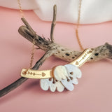 A handcrafted gold Tulip Necklace with engraved details rests on textured driftwood, featuring a white mother-of-pearl floral pendant, all set against a soft pink background for a touch of romantic charm.