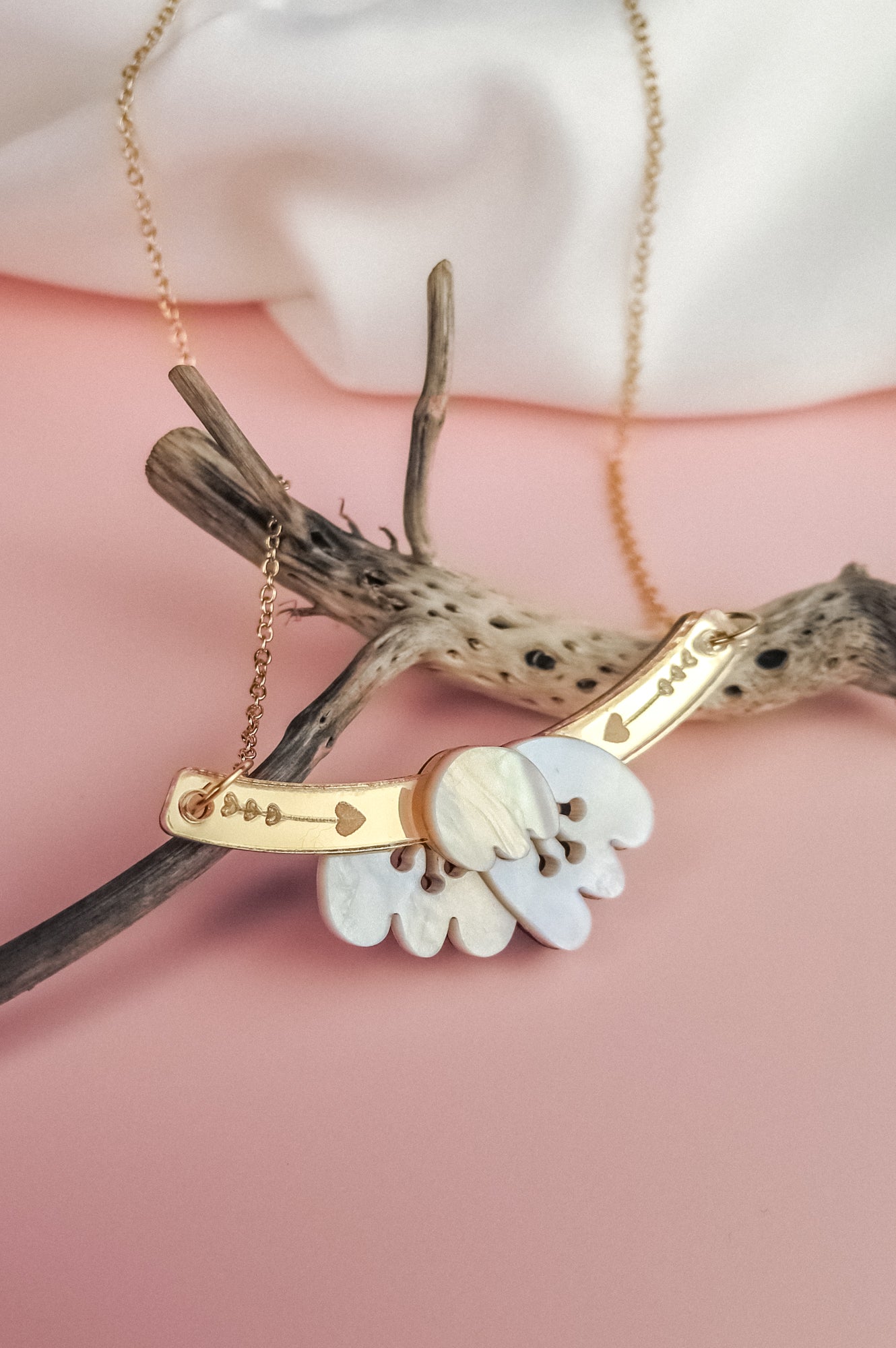 A handcrafted gold Tulip Necklace with engraved details rests on textured driftwood, featuring a white mother-of-pearl floral pendant, all set against a soft pink background for a touch of romantic charm.