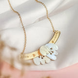 A gold Tulip Necklace with a delicate chain, featuring an intricate pendant with white and gold elements and small heart motifs, exudes romantic charm as it rests on a soft white fabric background.