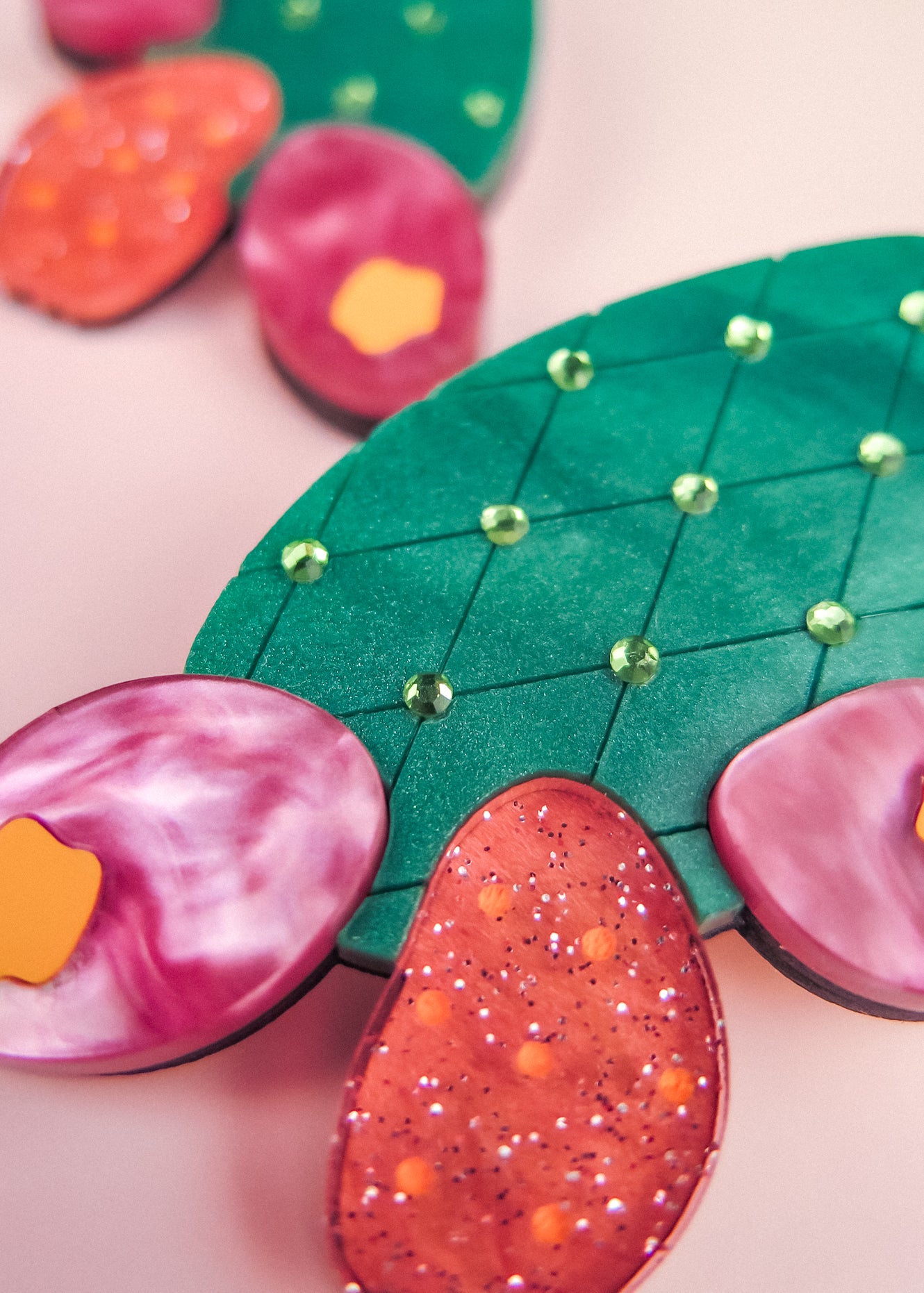 Brightly colored, stylized opuncia cactus artwork featuring a green pad with a diamond pattern and small gems, surrounded by pink and orange glittery and marbled pads—this bold design pops against a soft pink background.