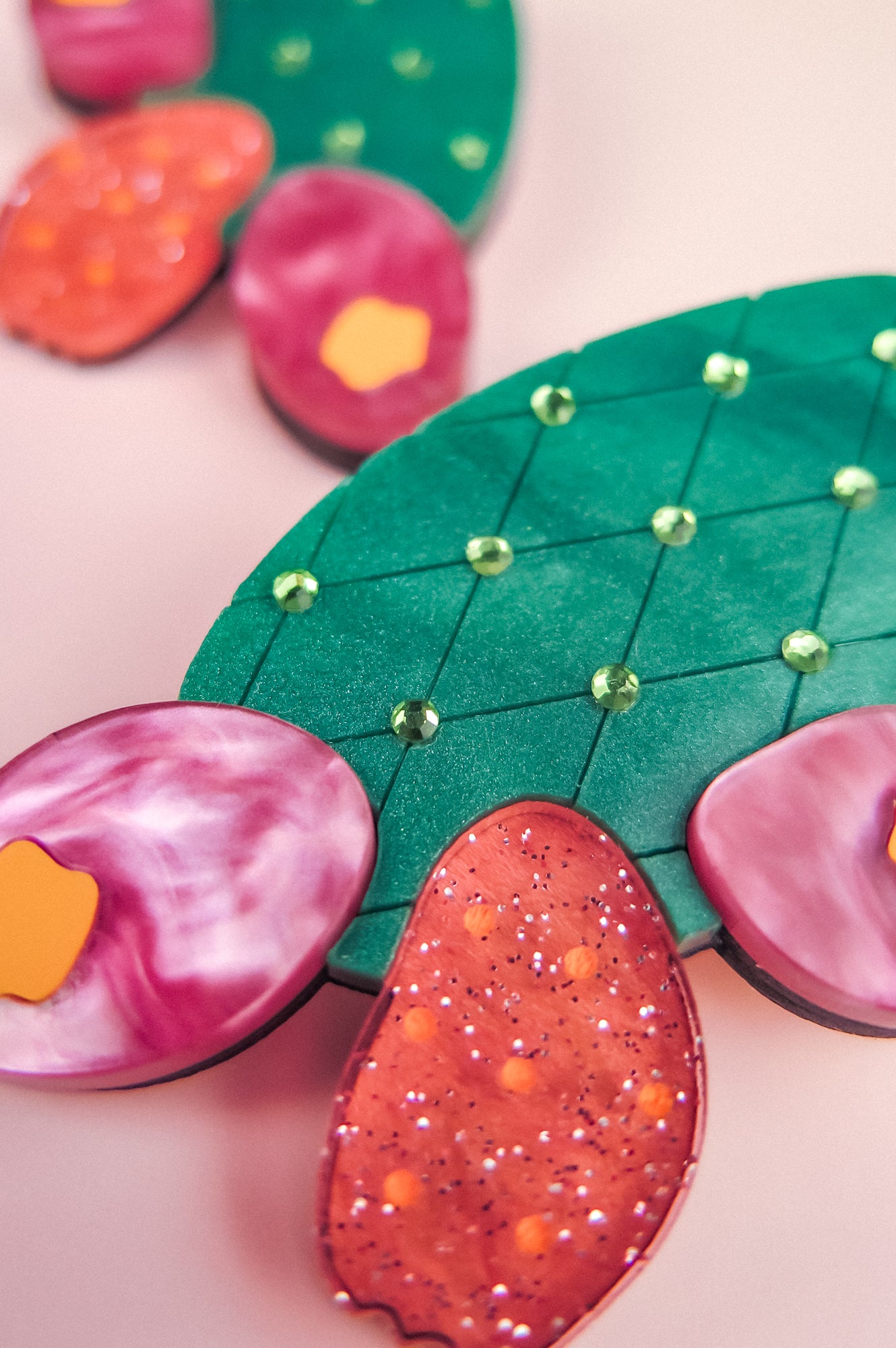 Brightly colored, stylized opuncia cactus artwork featuring a green pad with a diamond pattern and small gems, surrounded by pink and orange glittery and marbled pads—this bold design pops against a soft pink background.