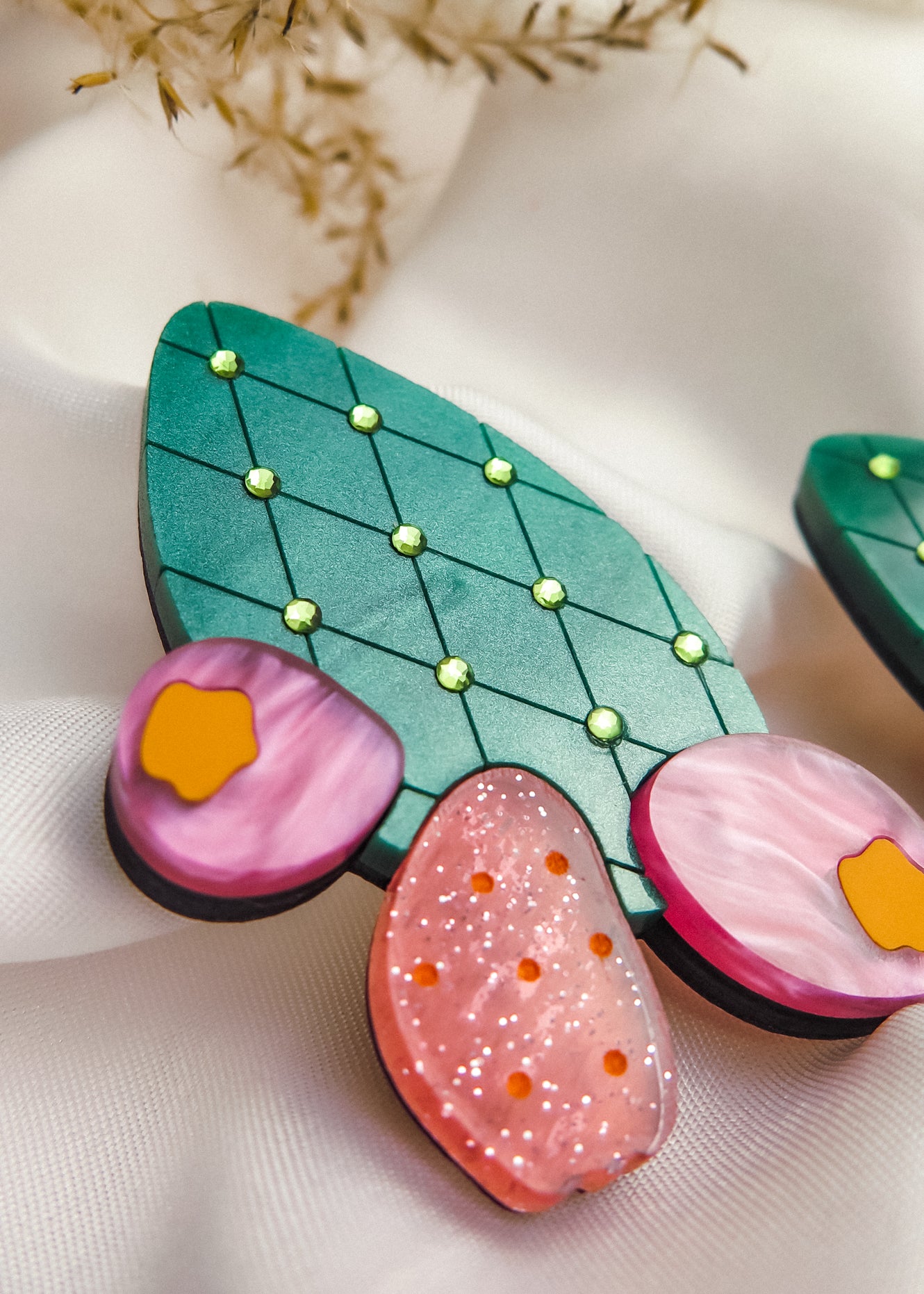 A close-up of bold statement earrings, stylized as green leaves with yellow dots and pink, orange, and purple accents—reminiscent of the opuncia cactus—displayed on soft white fabric with delicate dried flowers above.