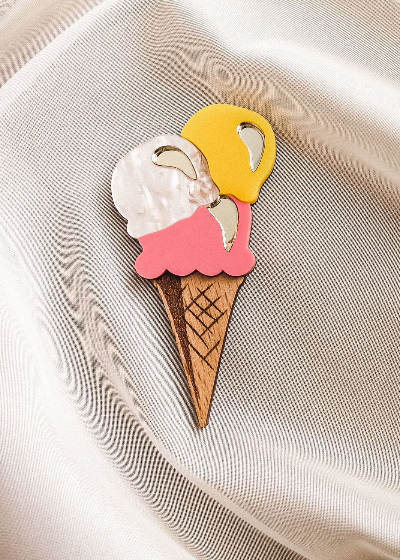 A handcrafted accessory, this Ice Cream Brooch features a wooden cone with yellow, white, and pink scoops, resting on smooth cream-colored satin—perfect for adding charm to your summer look.