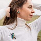 A woman in a white turtleneck stands outdoors with her hands in her hair, wearing matching colorful kingfisher bird earrings and a handcrafted Kingfisher Brooch. The background is a grassy field under a cloudy sky.