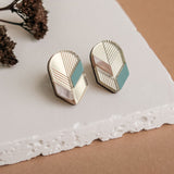 Handmade geometric earrings with gold, white, beige, and teal sections displayed on a white textured surface, dried flowers in the background—perfect for lovers of minimalist jewelry.