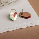 Two geometric earrings rest on a white textured surface. One showcases a modern design with gold lines and colorful accents, highlighting handmade minimalist jewelry; the other reveals its wooden and metal backing. Dried plant stems are in the background.