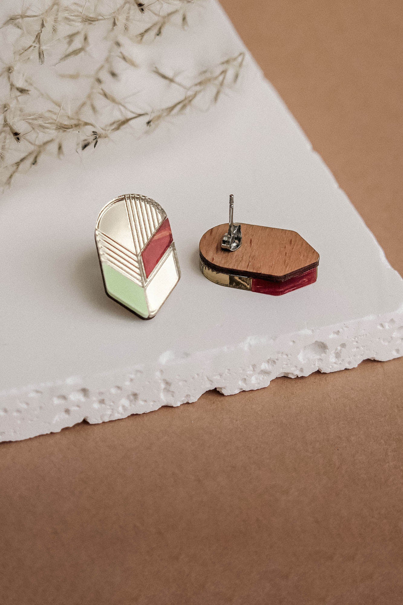 Two geometric earrings rest on a white textured surface. One showcases a modern design with gold lines and colorful accents, highlighting handmade minimalist jewelry; the other reveals its wooden and metal backing. Dried plant stems are in the background.