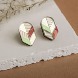 A pair of handmade geometric stud earrings with cream, mint green, and rust red sections outlined in silver, displayed on a textured white stone surface with wispy dried grass nearby—a perfect choice for minimalist jewelry lovers.
