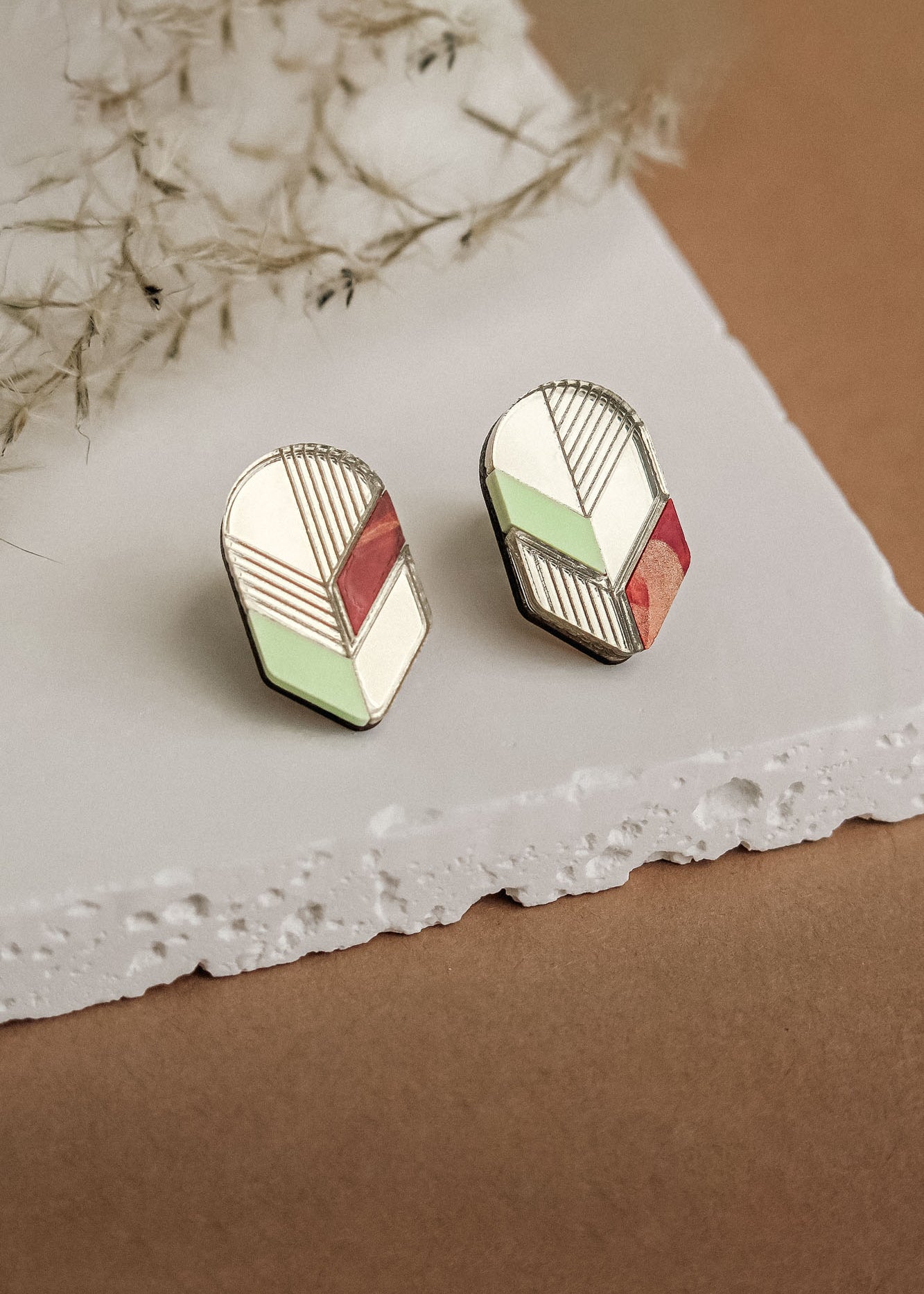A pair of handmade geometric stud earrings with cream, mint green, and rust red sections outlined in silver, displayed on a textured white stone surface with wispy dried grass nearby—a perfect choice for minimalist jewelry lovers.