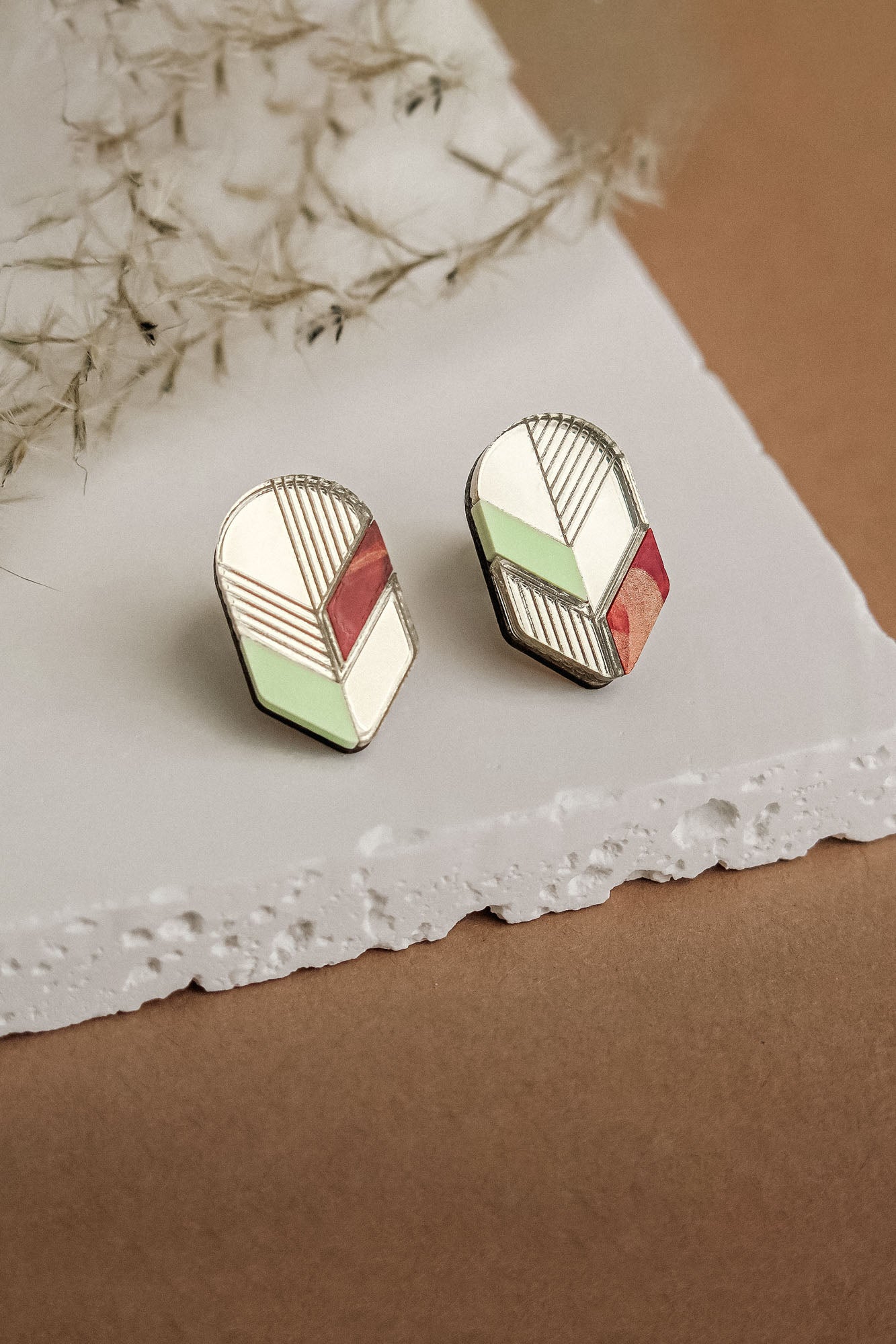 A pair of handmade geometric stud earrings with cream, mint green, and rust red sections outlined in silver, displayed on a textured white stone surface with wispy dried grass nearby—a perfect choice for minimalist jewelry lovers.