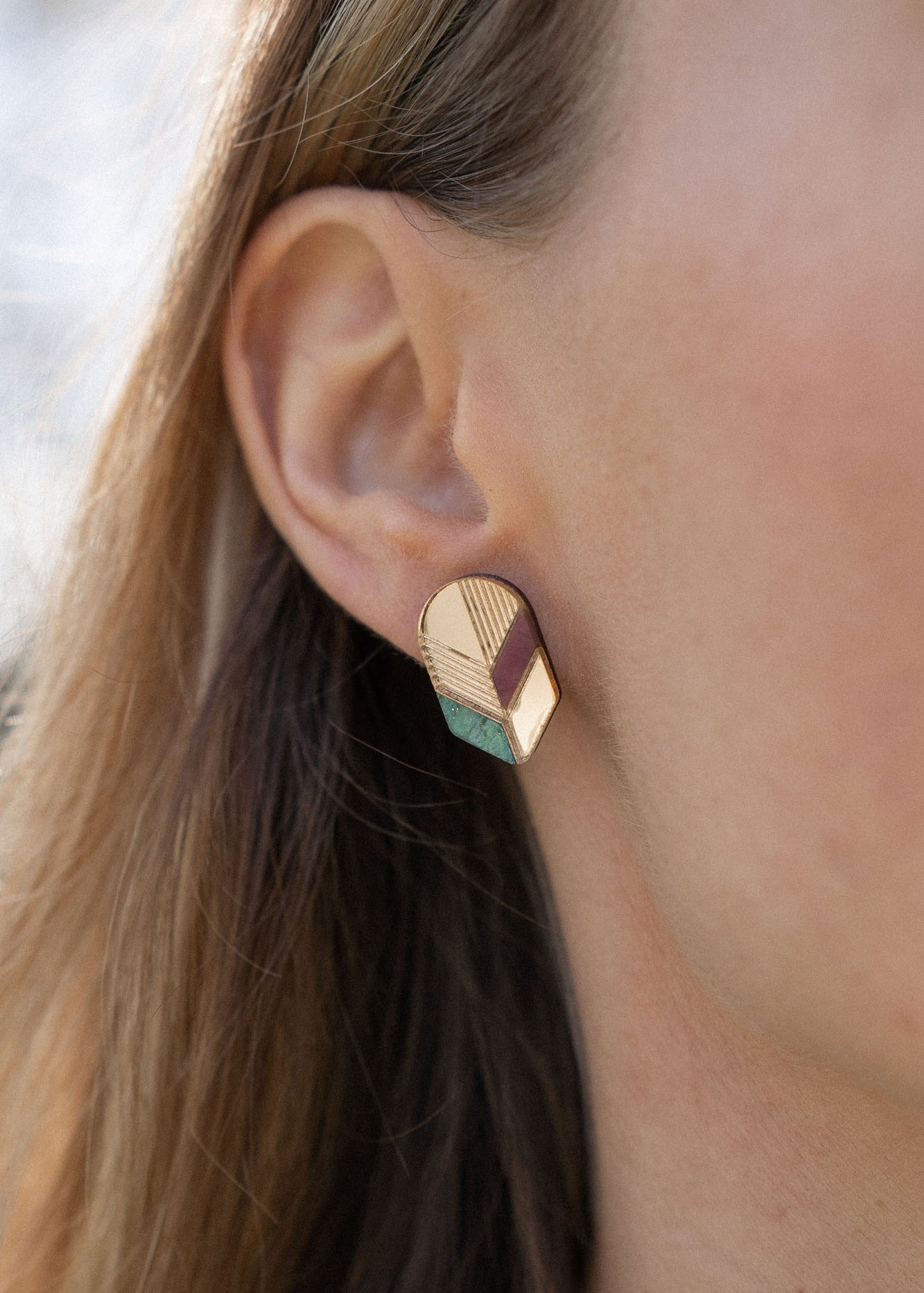 Close-up of a womans ear wearing a geometric, gold-toned stud earring with pastel accents. This piece of minimalist jewelry stands out against the softly blurred background, highlighting its unique handmade design and part of her hair and face.