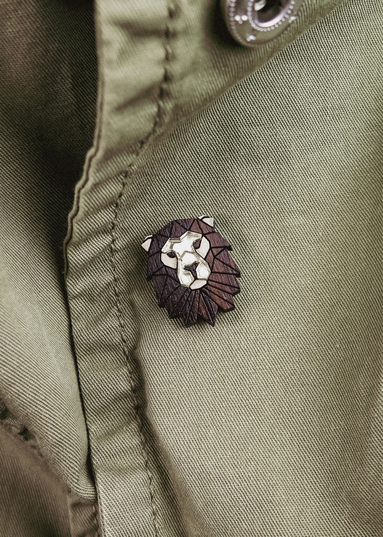 A close-up of an olive green jacket with a handcrafted geometric accessory—a Lion Pin—attached near a button.