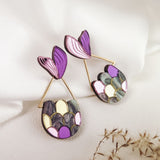 A pair of decorative earrings with purple, pink, gold, and gray patterns, shaped like abstract leaves and geometric forms, evokes whimsical elegance—perfect for mermaid lovers—displayed on white fabric with dried flowers nearby.