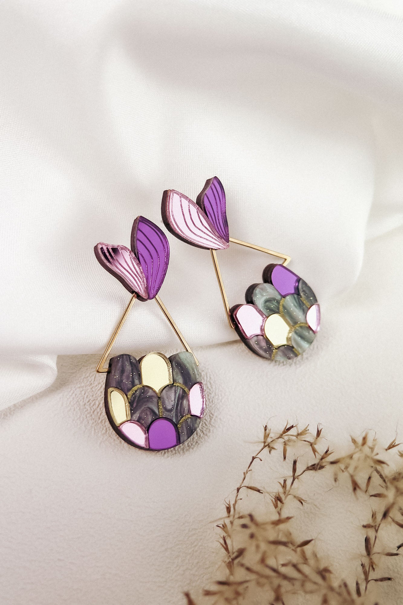 A pair of decorative earrings with purple, pink, gold, and gray patterns, shaped like abstract leaves and geometric forms, evokes whimsical elegance—perfect for mermaid lovers—displayed on white fabric with dried flowers nearby.